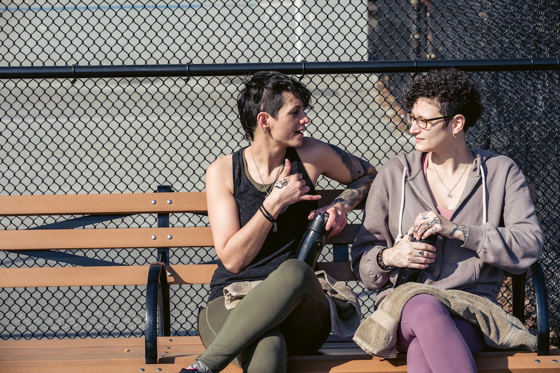 Comunicação Gestual. foto de Sarah Chai. Duas mulheres jovens sentadas num banco usando lingua de sinais